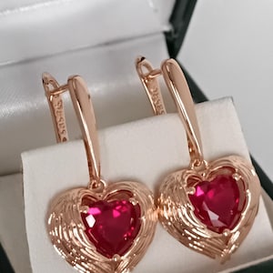 May include: A pair of rose gold heart-shaped earrings with a vibrant red gemstone center. The earrings feature a detailed, layered design around the heart, and are displayed in a jewelry box.