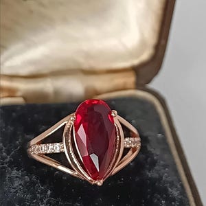 Op de afbeelding: Een roségouden ring met een grote, peervormige, dieprode gefacetteerde edelsteen als middelpunt. De ringband is aan beide kanten gespleten, waarbij de binnenste delen zijn versierd met kleine, heldere, fonkelende steentjes in het metaal.