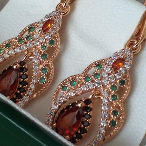 Puede incluir: Un par de pendientes de oro con una piedra roja en forma de lágrima, rodeada de piedras blancas y verdes más pequeñas. Los pendientes tienen un diseño delicado e intrincado.