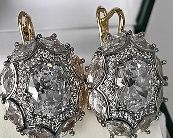 Puede incluir: Un par de pendientes dorados con una gran piedra central transparente de corte ovalado, rodeada por un halo de piedras transparentes más pequeñas de corte marquesa y un borde de diminutas piedras pavé, montados en una estructura plateada con cierre.