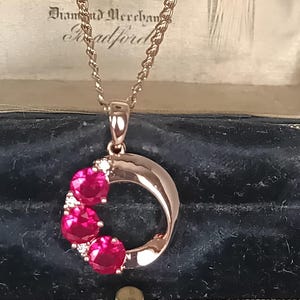 Op de afbeelding: Een roségouden ketting met een hanger in de vorm van een open cirkel, versierd met drie ronde, felrode stenen en kleine heldere accentstenen. De hanger hangt aan een fijne roségouden ketting met een gepolijste metalen afwerking.