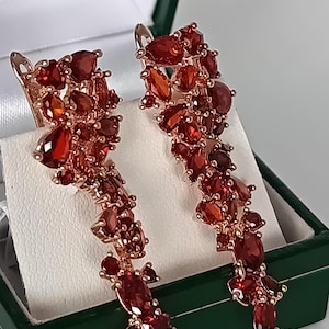 May include: A pair of long drop earrings featuring clusters of faceted, deep red, pear-shaped and oval-cut stones set in a rose-gold tone metal. The earrings are displayed vertically, showcasing a cascading arrangement of stones from top to bottom.