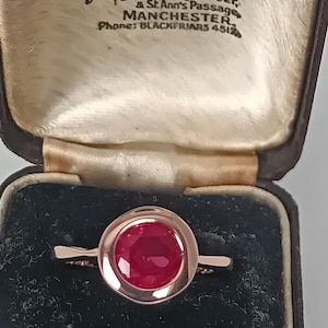 Könnte beinhalten: Ein Ring mit einem runden, facettierten, dunkelrosa Edelstein in einer glatten, polierten Metallschiene. Der Ring befindet sich in einer dunklen, aufklappbaren Schmuckschatulle mit einem cremefarbenen Innendeckel, der den Text Hancock & Son Ltd. trägt.