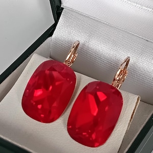 Puede incluir: Un par de pendientes de cristal rojo rectangulares con ganchos dorados. Los pendientes se exhiben en una caja de joyería negra con un forro de satén blanco. Los cristales tienen múltiples facetas.