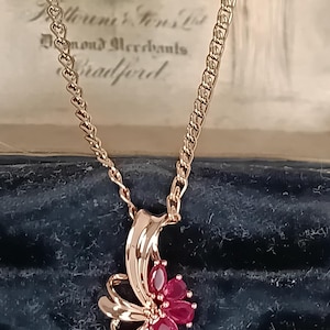 May include: A rose gold-toned necklace featuring a pendant with a polished, curved metal ribbon design and a cluster of five marquise-cut, deep red gemstones arranged in a floral pattern, suspended from a delicate, matching rose gold-toned chain.