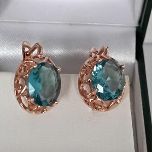 May include: A pair of rose gold earrings with large, round, teal-colored gemstones. The earrings have an ornate, openwork design around the gemstones, and are displayed in a white jewelry box.