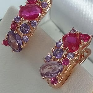 Peut inclure: Une paire de boucles d'oreilles dorées ornées de grappes de pierres précieuses roses et violettes. Les boucles d'oreilles ont une fermeture à levier et sont serties de pierres de taille ovale et ronde. Les pierres sont disposées en motif floral.
