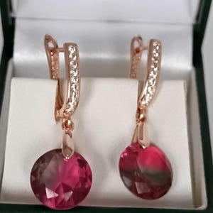 Könnte beinhalten: Ohrringe aus Roségold mit einer Reihe kleiner, klarer Steine am Ohrdraht und einem großen, runden, rosa- und dunkelroten Kristallanhänger. Die Ohrringe werden in einer grünen Schmuckschatulle präsentiert.