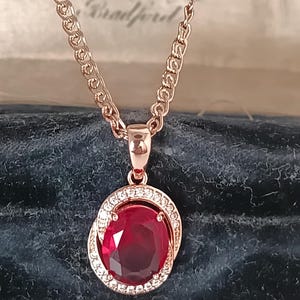 Pode incluir: Colar com pingente em ouro rosa com uma grande gema de rubi oval. O rubi é rodeado por pequenos cristais transparentes. A corrente é em ouro rosa. O colar é apresentado sobre uma superfície de veludo azul escuro. O rubi do pingente tem cerca de 2 cm de comprimento.
