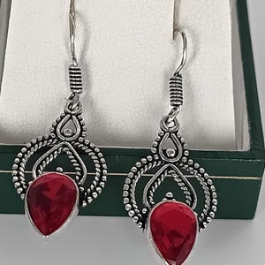May include: Silver-tone drop earrings featuring a teardrop-shaped, faceted red gemstone. The earrings have an ornate, openwork design with a spiral detail near the hook. The earrings are displayed in a green jewellery box.
