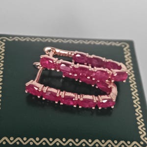 May include: Rose gold hoop earrings, each set with a line of vivid, oval-cut rubies. The earrings are presented on a dark green jewellery box with a decorative border.