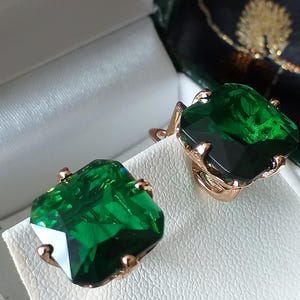 Könnte beinhalten: Ein Paar goldene Ohrringe mit großen, quadratischen, smaragdgrünen Edelsteinen. Die Ohrringe befinden sich in einer weißen Schachtel mit grüner Auskleidung.
