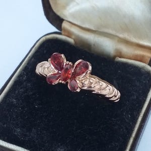 Könnte beinhalten: Ein roségoldener Ring mit Schmetterlingsdesign, besetzt mit roten Edelsteinen. Der Ring wird in einer Vintage-Schmuckschatulle mit schwarzem Samt präsentiert. Die Schatulle hat Text.