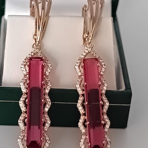 Op de afbeelding: Een paar lange oorbellen met een rechthoekige, dieproze edelsteen in een roségouden zetting. Het frame is omrand met een golvende, geschulpte rand die is ingelegd met kleine, heldere witte steentjes en heeft een stevige sluiting aan de achterkant.