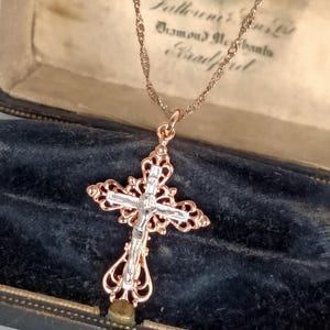 Könnte beinhalten: Eine Halskette mit Kreuzanhänger aus Roségold mit einer silbernen Jesusfigur. Das Kreuz hat ein verziertes Filigran-Design. Die Halskette wird in einer Vintage-Schmuckschatulle mit Text präsentiert.
