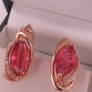 Könnte beinhalten: Ein Paar roségoldene Ohrringe mit großen, oval geformten, rosa Edelsteinen. Die Ohrringe haben ein dekoratives Design mit kleinen, klaren Steinen um den rosa Edelstein. Die Ohrringe werden in einer weißen Schmuckschatulle präsentiert.