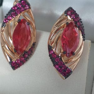 May include: A pair of gold-coloured earrings with a leaf-like design. Each earring features a large, oval, red gemstone in the centre, surrounded by smaller, pink gemstones and black accents. The earrings are displayed in a jewellery box.
