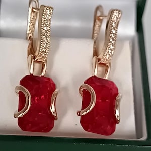 Könnte beinhalten: Ein Paar goldfarbene Ohrringe mit roten rechteckigen Edelsteinen. Der obere Teil der Ohrringe hat ein gebogenes Design mit kleinen, klaren Steinen. Die Ohrringe werden in einer grün-weißen Schmuckschatulle präsentiert.