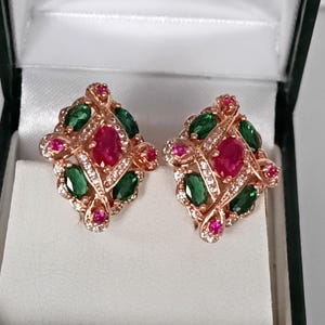 Pode incluir: Brincos em ouro rosa com um rubi oval central, rodeado por pedras preciosas verdes e rosas e pequenos cristais transparentes. Os brincos são apresentados numa caixa de joias a preto e branco.