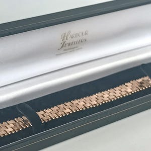 May include: A rose gold bracelet with a geometric design rests in a black jewelry box with a white interior. The box is open, revealing the bracelet's intricate pattern. The box is labeled "Harbour Jewellers".