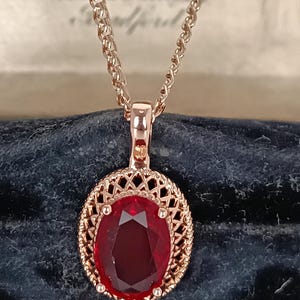 May include: Rose gold pendant necklace featuring an oval, deep red gemstone. The gemstone is set within a decorative, openwork frame. The chain is a delicate, linked design. The necklace is displayed against a dark blue background.