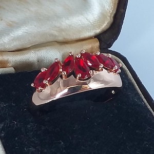 Könnte beinhalten: Ein roségoldener Ring mit einer Reihe von tiefroten, marquiseförmigen Edelsteinen. Der Ring ist in einem polierten Band gefasst und wird in einer Vintage-Schmuckschatulle präsentiert. Ein klassisches Schmuckstück.