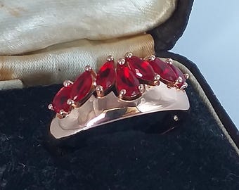 May include: A rose gold ring featuring a series of marquise-cut, deep red gemstones. The ring is set in a polished band and is displayed in a vintage jewelry box. The ring is a classic piece of jewelry.