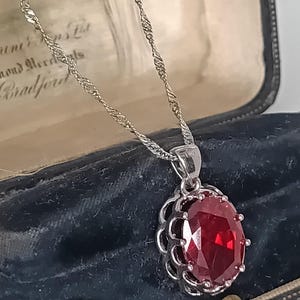 Könnte beinhalten: Eine silberfarbene Halskette mit einem großen, oval geschliffenen, tiefroten Edelsteinanhänger in einer dekorativen, gewellten Metallfassung. Der Anhänger hängt an einer zarten, gedrehten silberfarbenen Kette und hat eine facettierte Oberfläche.