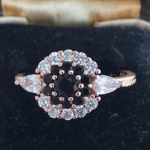 Può includere: Anello in oro rosa con una pietra nera centrale circondata da pietre bianche e nere. Due pietre bianche a forma di pera affiancano il design centrale. L'anello è posto su uno sfondo scuro.