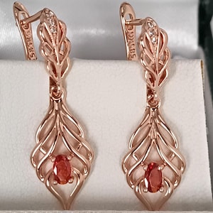 Puede incluir: Un par de pendientes de oro rosa con un intrincado diseño de plumas. Cada pendiente presenta una piedra preciosa naranja de corte ovalado y pequeñas piedras de acento. Los pendientes se exhiben en una caja blanca con un borde verde.