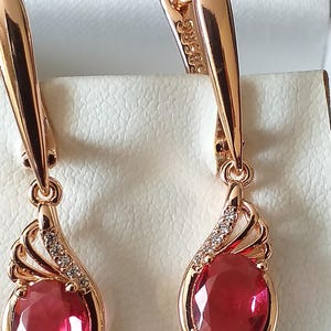 Puede incluir: Un par de pendientes de oro con gotas de piedras preciosas rojas y pequeñas piedras transparentes. Los pendientes tienen un diseño delicado y femenino.
