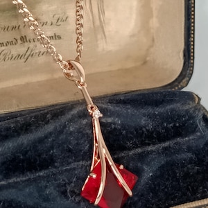Op de afbeelding: Een roségouden hangerketting met een geometrisch ontwerp. De hanger heeft een rode, ruitvormige edelsteen en een kleine, heldere accentsteen. De ketting wordt gepresenteerd in een vintage sieradendoosje.