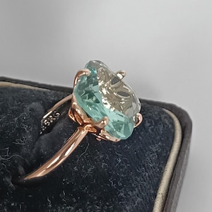 Puede incluir: Un anillo de oro rosa con una gran piedra preciosa azul claro de corte irregular. La piedra preciosa está sujeta por un engaste detallado. La banda del anillo es lisa y pulida. El anillo se muestra sobre una superficie de terciopelo azul oscuro.