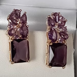 Op de afbeelding: Een paar goudkleurige oorbellen. Elke oorbel heeft een rechthoekige, donkerpaarse edelsteen aan de basis, bekroond met een cluster van kleinere, druppelvormige en ronde paarse edelstenen. Op een witte achtergrond.