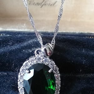 Pode incluir: Um colar de corrente de prata com um pingente que apresenta uma grande gema verde ovalada cercada por um halo de gemas transparentes menores.