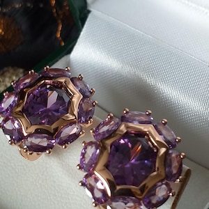 Peut inclure: Une paire de boucles d'oreilles en améthyste avec un sertissage en or rose, présentée dans un écrin doublé de satin blanc. Chaque boucle d'oreille présente une grosse pierre violette centrale entourée de pierres ovales plus petites.
