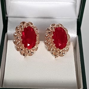 May include: A pair of rose gold-coloured stud earrings featuring large, oval, red gemstones. The earrings are set in a decorative, scalloped frame and are presented in a green and white jewellery box.
