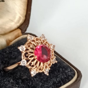 Op de afbeelding: Een roségouden ring met een centrale, rond geslepen, felroze edelsteen omringd door een decoratieve filigraan metalen zetting. De buitenrand van het bloemvormige ontwerp is geaccentueerd met acht kleine, heldere, rond geslepen stenen in pootjes.