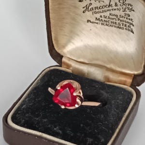 Könnte beinhalten: Ein roségoldener Ring mit einem herzförmigen, roten Edelstein, präsentiert in einer Vintage-Schmuckschatulle in Braun. Die Schatulle ist mit cremefarbenem Satin ausgekleidet und trägt den Text "Hancock & Son".