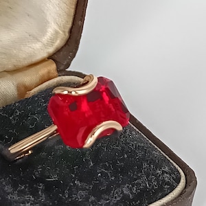 Op de afbeelding: Een vintage ring met een grote, gefacetteerde rode edelsteen gezet in goudkleurig metaal. De ring wordt gepresenteerd in een bruine sieradendoos met een zwart fluwelen interieur. De edelsteen heeft een onregelmatige vorm.