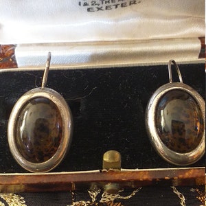 Pode incluir: Um par de brincos de prata com pedras marrons ovais. Os brincos estão em uma caixa de madeira com um forro de cetim branco. A caixa tem um rótulo que diz "Goldsmiths & Jewellers, 1 & 2 The Arcade, Exeter."