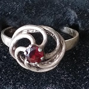 May include: A silver ring featuring a swirling design, encircling a central, round, deep red gemstone. The ring is set against a dark, textured backdrop, possibly a jewellery display.