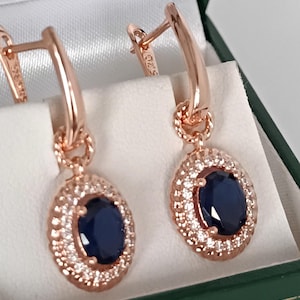 May include: Rose gold earrings featuring oval, dark blue gemstones encircled by small, clear stones. The earrings are presented in a green jewellery box with a white interior. The earrings have a lever back closure.