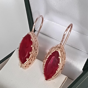 May include: A pair of rose gold earrings featuring large, oval-cut, deep red gemstones. The earrings are set in a decorative frame and are presented in a green and white jewellery box. The earrings have a lever back closure.