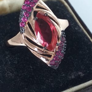 Könnte beinhalten: Roségoldener Ring mit einem großen, oval geschliffenen, roten Edelstein im Zentrum. Der Ring ist mit kleineren, runden, roten Edelsteinen und schwarzen Akzenten verziert. Der Ring befindet sich in einer schwarzen Schmuckschatulle.