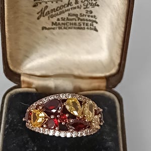 Puede incluir: Un anillo con piedras preciosas rojas y amarillas en talla pera y marquesa, montado en una banda con pequeñas piedras claras. El anillo se presenta en un joyero marrón con el texto "Hancock & Son (Goldsmiths) Ltd" impreso en el interior.