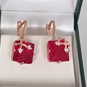 Könnte beinhalten: Ein Paar roségoldene Ohrringe mit quadratischen, tiefroten Edelsteinen. Jeder Ohrring hat einen kleinen, funkelnden Akzentstein und ein kreuzförmiges Design. Die Ohrringe werden in einer grün-weißen Schmuckschatulle präsentiert.