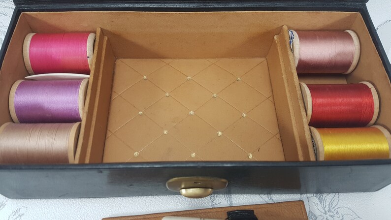 May include: An open black box with a gold latch. The box is lined with brown fabric and contains spools of thread in various colors, including pink, purple, red, yellow, and beige.