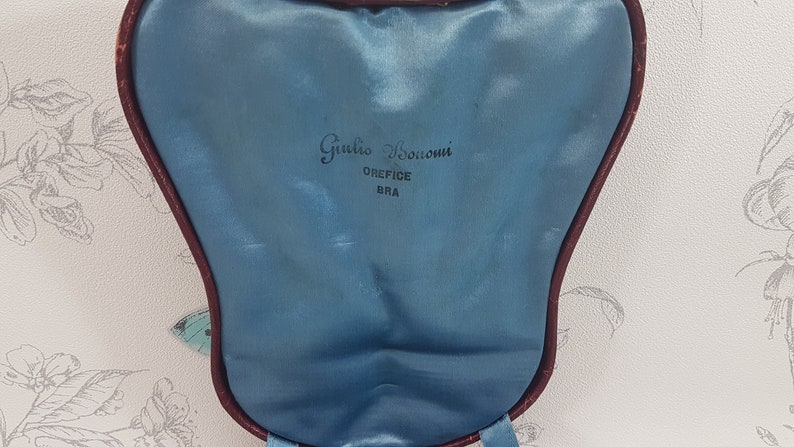 May include: A blue velvet jewellery box with a burgundy trim. The box has the text "Giulio Bocconi" and "OREFICE BRA" printed on the inside.