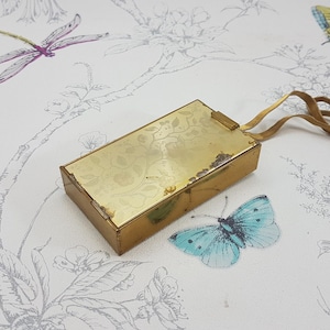 May include: A gold-colored compact with a floral design and a rubber strap. The compact is closed and sitting on a white surface with a floral pattern.